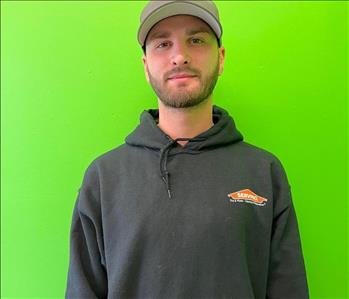 man with a grey SERVPRO sweatshirt and a hat standing in front of a green background. 