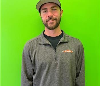 man in a grey SERVPRO shirt and hat standing in front of a green background. 